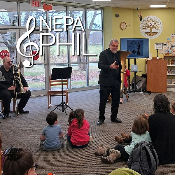 This image captures a community music education event, likely hosted by the NEPA Philharmonic (logo displayed in the upper left). A man in black, possibly a conductor or musician, is speaking to a group of young children seated on the floor, engaging them in learning about music. Another musician with a trombone sits nearby, while a tuba is also present. Behind them, a screen displays the words “Bent Trumpet” alongside an image, suggesting a playful or educational music program. The setting appears to be a public library, emphasizing accessibility and outreach.