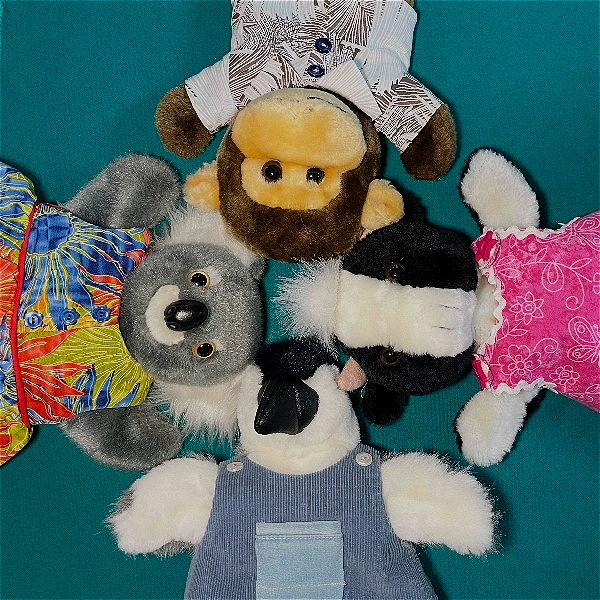 This image shows four plush toys arranged in a circle, lying on a teal surface with their heads pointing toward the center. Each stuffed animal is dressed in colorful clothing: A brown monkey in a patterned shirt (top). A gray koala in a bright Hawaiian-style shirt (left). A black-and-white cow in denim overalls (bottom). A white dog with black patches in a pink dress (right). They look like they’re having a playful "meeting" or group huddle.