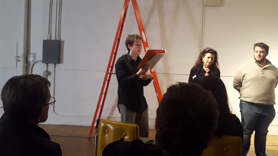 This photo shows a stage reading of a play. One performer is reading from a book to their left or two other performers. We can also see some audience members in the four ground looking towards them.