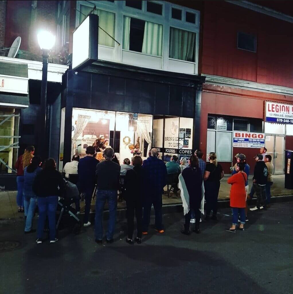 This photo shows a lively street performance in a storefront window at night. A crowd has gathered outside a shop with large front windows, watching performers inside. The shop sign reads “Bring Ideas to Life” and mentions folders and copies, suggesting it’s usually a print or copy store. People stand and sit, filling the sidewalk, creating a casual, community-centered atmosphere. The audience is wearing headphones to hear the audio of the performance.