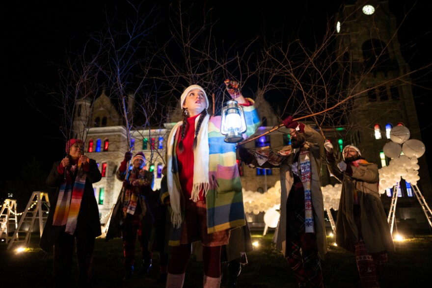 A photo of a female holding a lantern she is dressed in a red sweater with a jacket of horizontal rainbow striped colors. She is wearing a knit hat and looking upwards into the sky. She is outdoors and standing in front of four other performers in the background, also in winter attire, there is a building behind them in the distance.