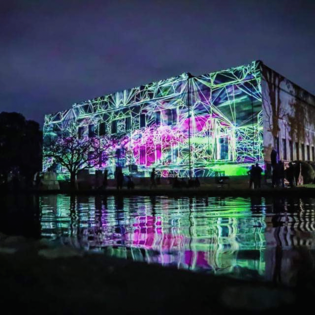 This image shows a nighttime projection mapping display on the side of a large building. Neon-colored geometric lines and shapes in green, purple, and pink create a glowing web-like effect across the structure. The vivid visuals reflect beautifully in a body of water in the foreground, enhancing the surreal atmosphere.