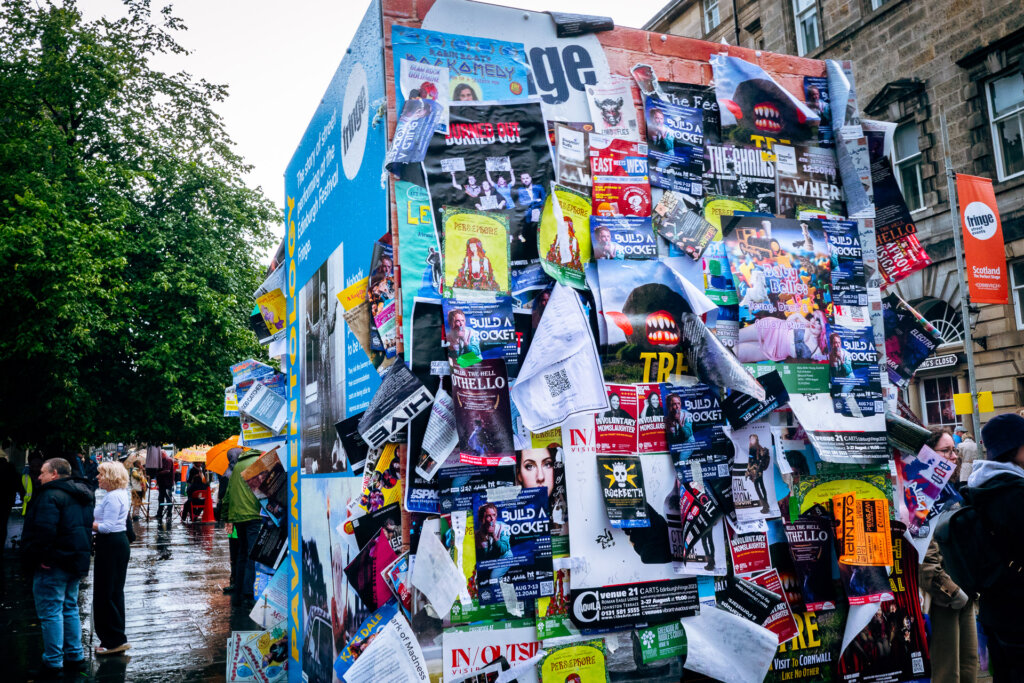 This photo shows a display of many different posters at the Edinburgh Fringe. It appears to be raining.