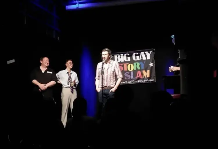 This image shows a live storytelling event with four people on stage in front of an audience. A man at the center stands at a microphone, speaking, while two others to his left smile and watch. Behind them, a banner reads “BIG GAY STORY SLAM – A celebration of LGBTQ voices and allies”, with the words “Story Slam” in colorful rainbow letters. The stage is dimly lit with blue and purple spotlights, creating an intimate performance setting.