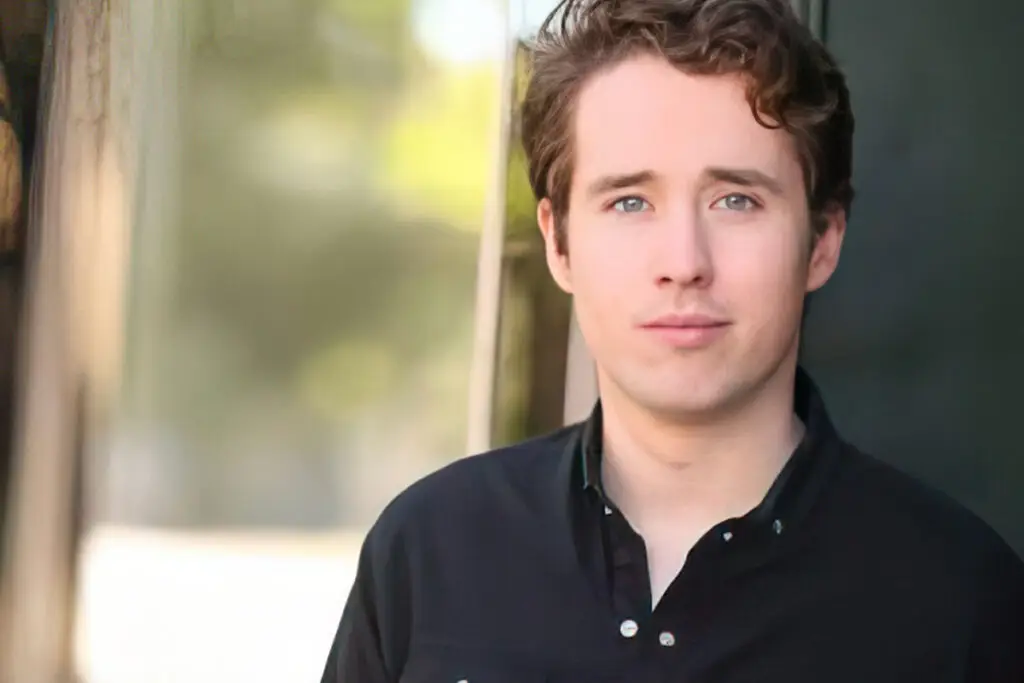 This image shows Conor O'Brien with short, wavy brown hair and bright blue eyes, standing outdoors. He is wearing a black collared shirt with the top button open, giving him a relaxed but polished appearance. The background is softly blurred with shades of green and beige, keeping the focus on his face.