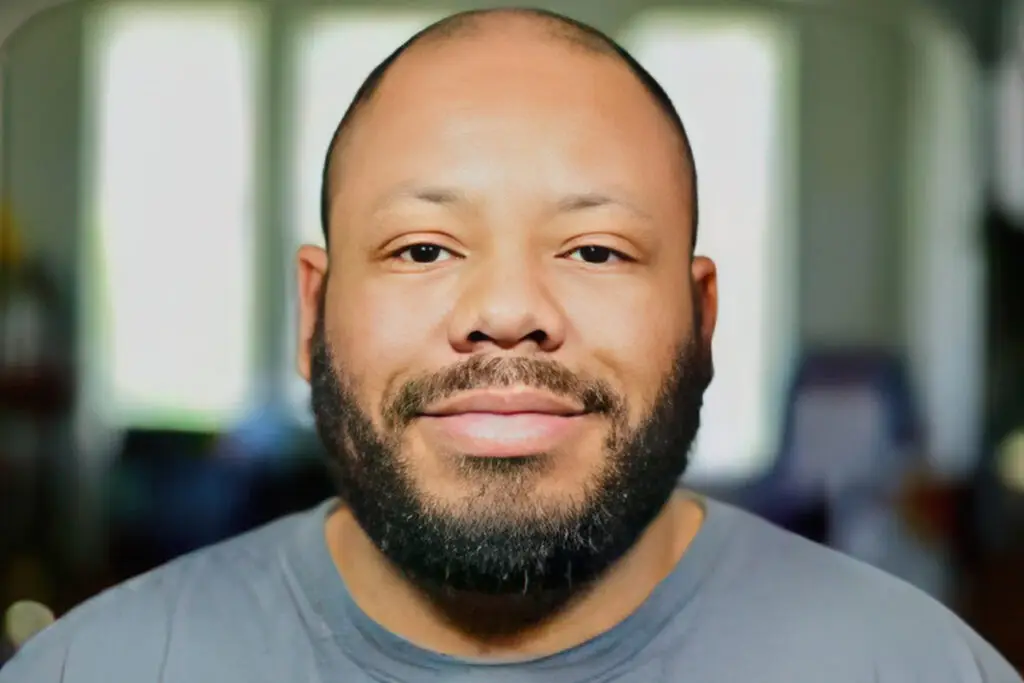 This image shows a close-up portrait of Dan Kimbrough with a neatly groomed beard and shaved head. He is wearing a casual gray shirt and has a calm, slight smile on his face. The background is softly blurred, making the focus entirely on his face.