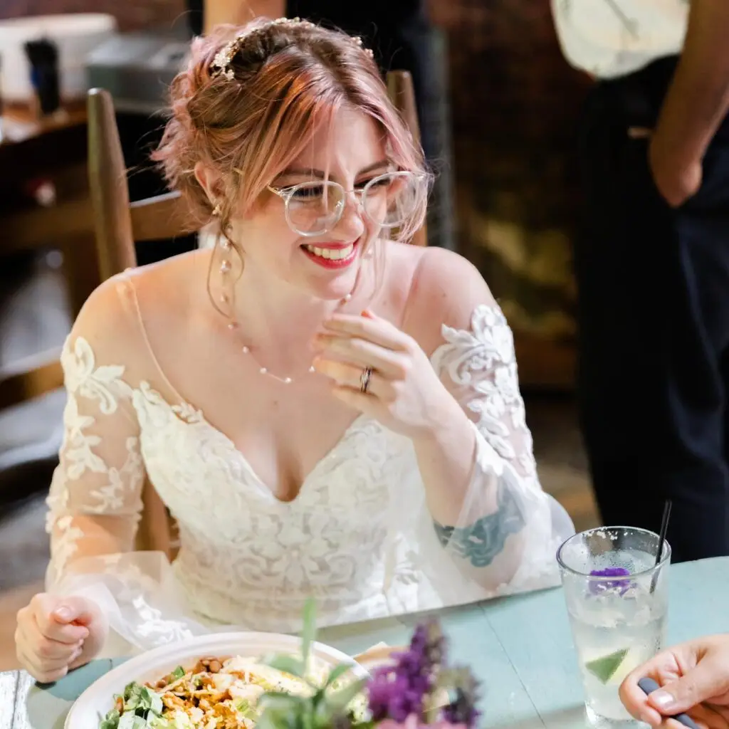 This image shows a smiling bride in a white lace wedding dress sitting at a table. She has light hair styled up with decorative hair accessories and is wearing glasses and a delicate necklace. In front of her is a plate of food and a glass with a drink, and she looks joyful, possibly during a wedding reception meal.