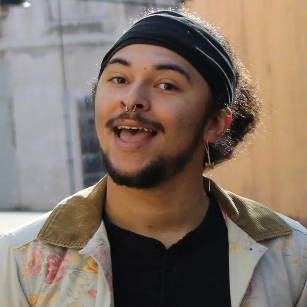 This image shows Terry Thompson smiling warmly at the camera, creating an open and friendly vibe. They are wearing a black headband, a septum piercing, and a long earring on one ear. Their outfit includes a floral-patterned jacket with a tan collar layered over a black shirt, giving a stylish and expressive appearance.