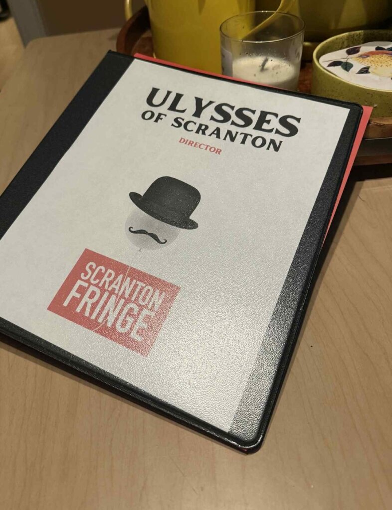 This photo shows a binder with the name Ulysses of Scranton in Black and the word director in red underneath the Scranton fringe logo is at the bottom and floating in front of it is a white balloon with a fake mustache and a bowler hat