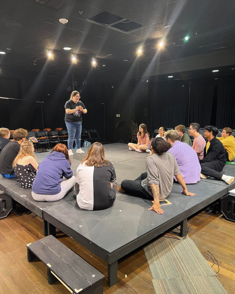 Photo of a group of performers sitting in a half circle on a raised stage. One person is standing and speaking to the group.