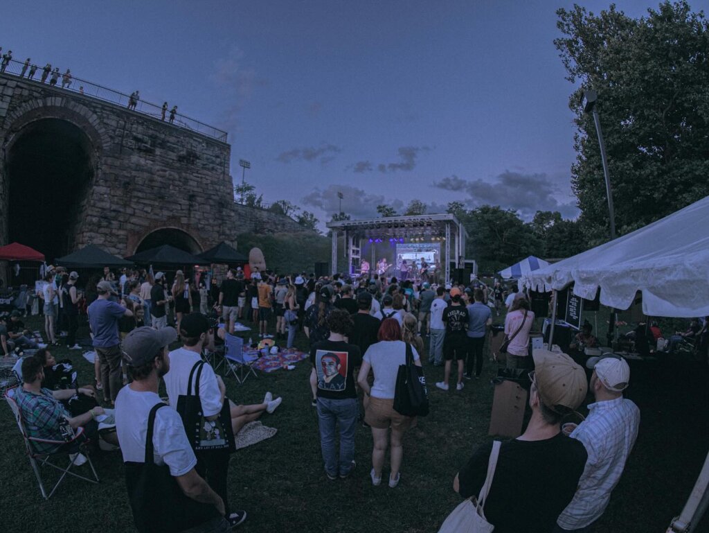 Photo of an outdoor concert in the evening. There is a crowd of people facing the large illuminated outdoor stage, there is a stone wall of a building to the left and the sky is a dark blue.