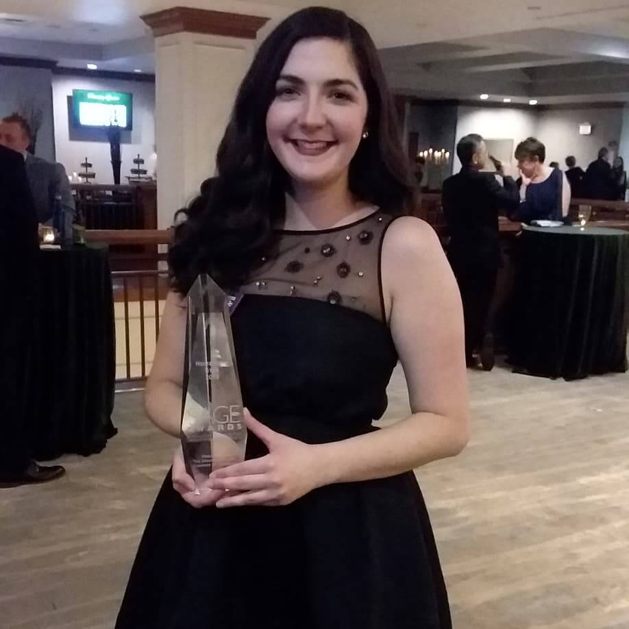Photo of a smiling woman with dark hair, wearing a black dress, holding a Sage Award in her hand.