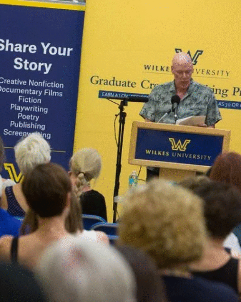 A older man with a bald head and wearing a flowered print shirt is standing behind a Wilkes University podium and reading to the audience. To the right is a banner that reads, "Share Your Story"