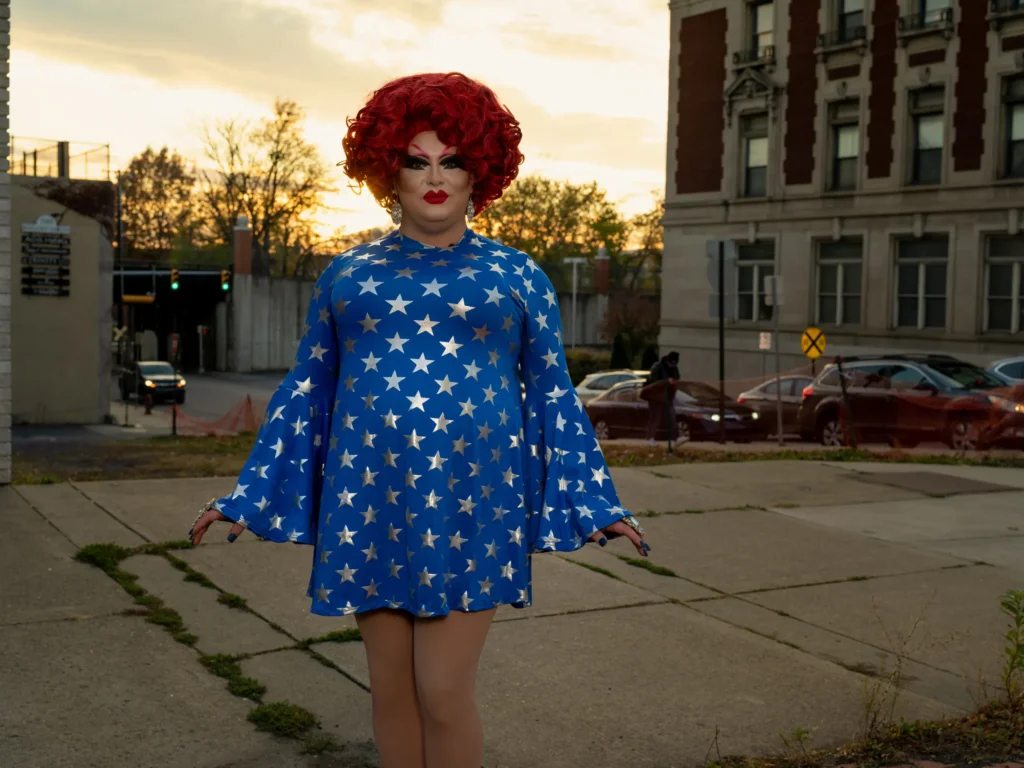 Photo of a drag queen with curly red hair and a dress of blue with silver stars. They are standing in an empty cement lot.