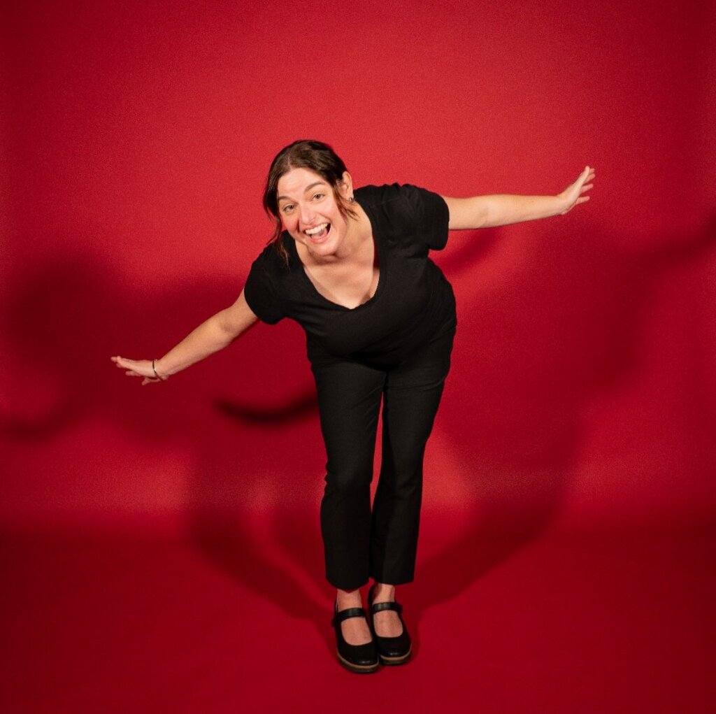 Photo of Mandy Pennington wearing all black clothing, bending down at the waist with her arms outstretched in a bow like position against a red background. 